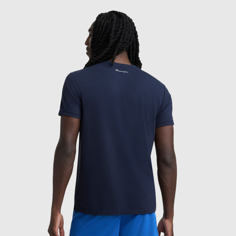 A person with long, twisted hair is seen from the back wearing a Champion Sport Crewneck T-Shirt, Script Logo in navy blue and bright blue lightweight micro mesh shorts against a plain light gray background.