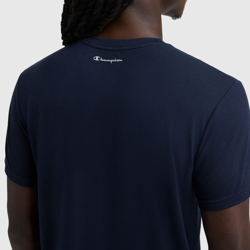 A person with dark skin and long hair is seen from the back wearing a navy blue Champion Sport Crewneck T-Shirt, Script Logo, with the Champion logo near the neckline.