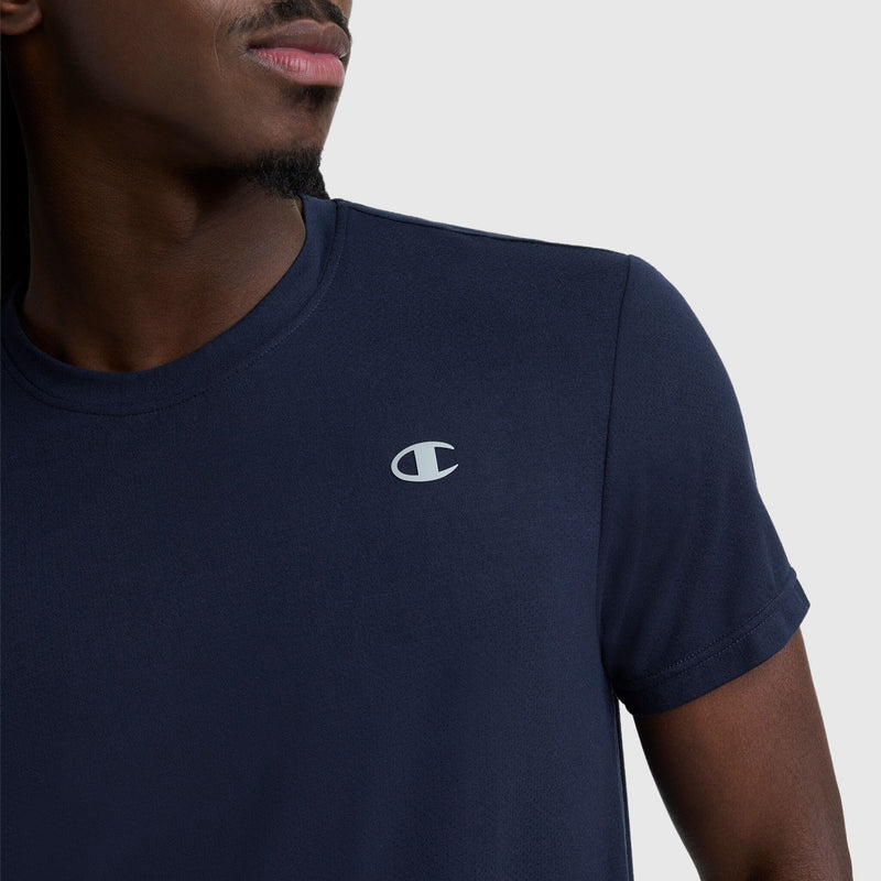 A person wearing a navy blue Champion Sport Crewneck T-Shirt, Script Logo, with the Champion logo on the left chest, shown from the shoulders up against a plain light background.