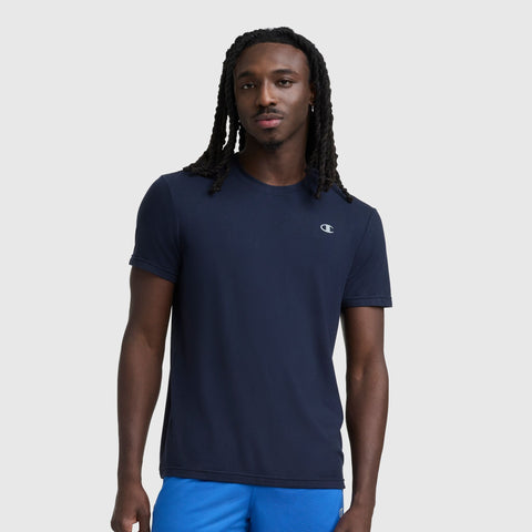 A man with long, braided hair wears a navy Sport Crewneck T-Shirt, Script Logo by Champion and bright blue shorts, standing against a plain light gray background.