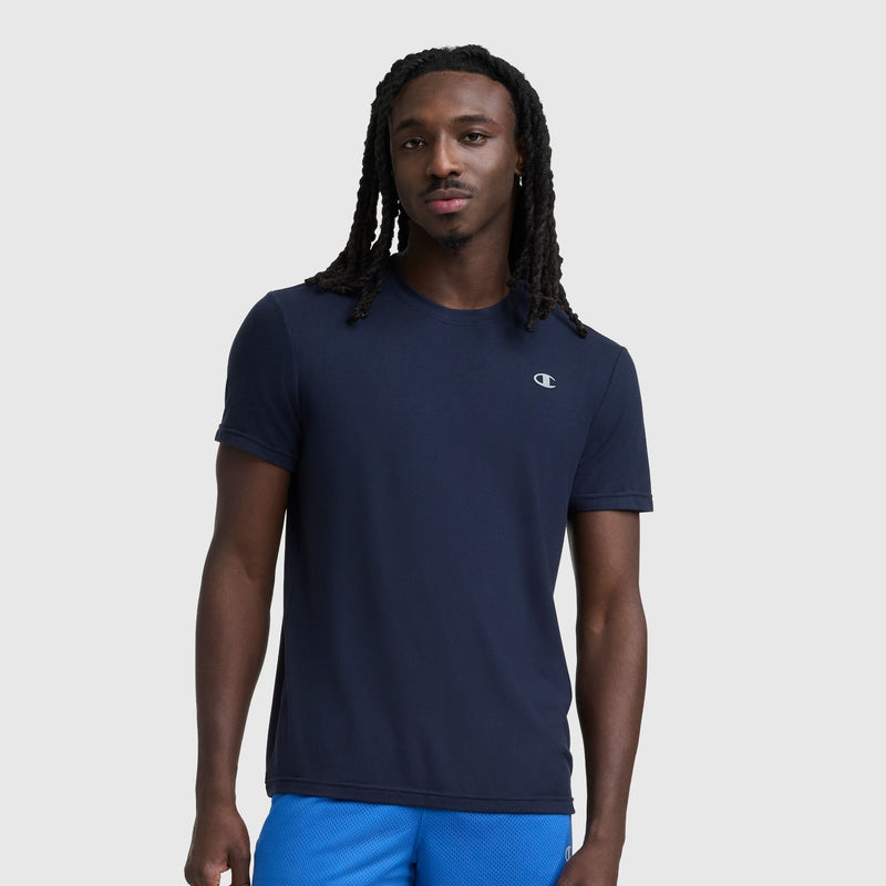 A man with long, braided hair wears a navy Sport Crewneck T-Shirt, Script Logo by Champion and bright blue shorts, standing against a plain light gray background.