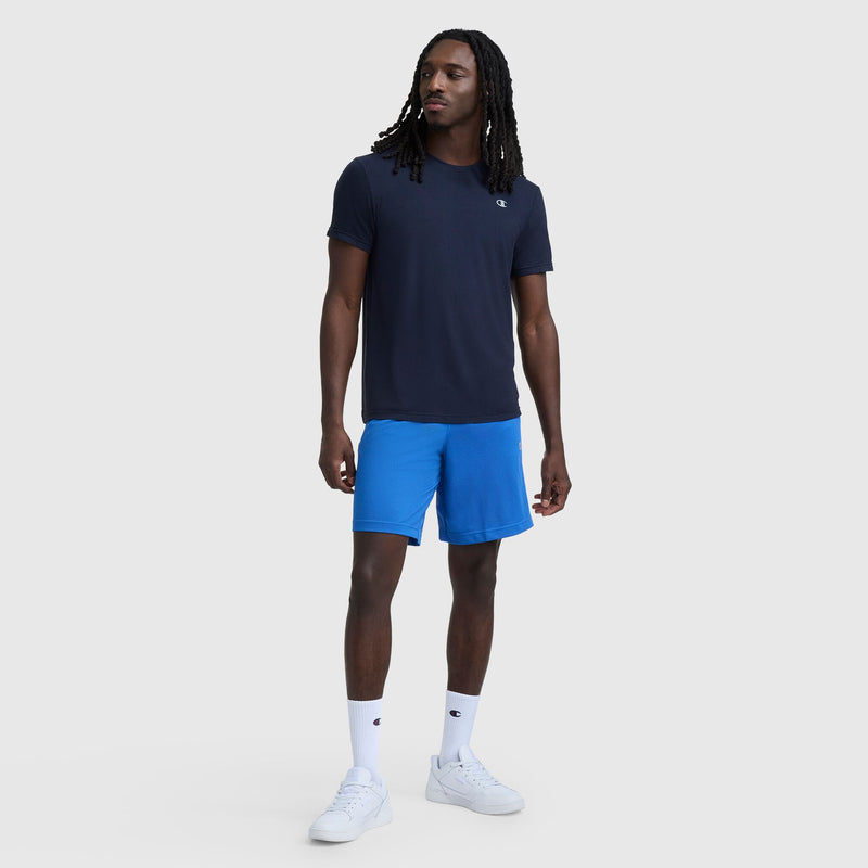 A man with long braids looks left, sporting a Champion Sport Crewneck T-Shirt with Script Logo in navy, bright blue athletic shorts, white crew socks, and white sneakers against a plain background.