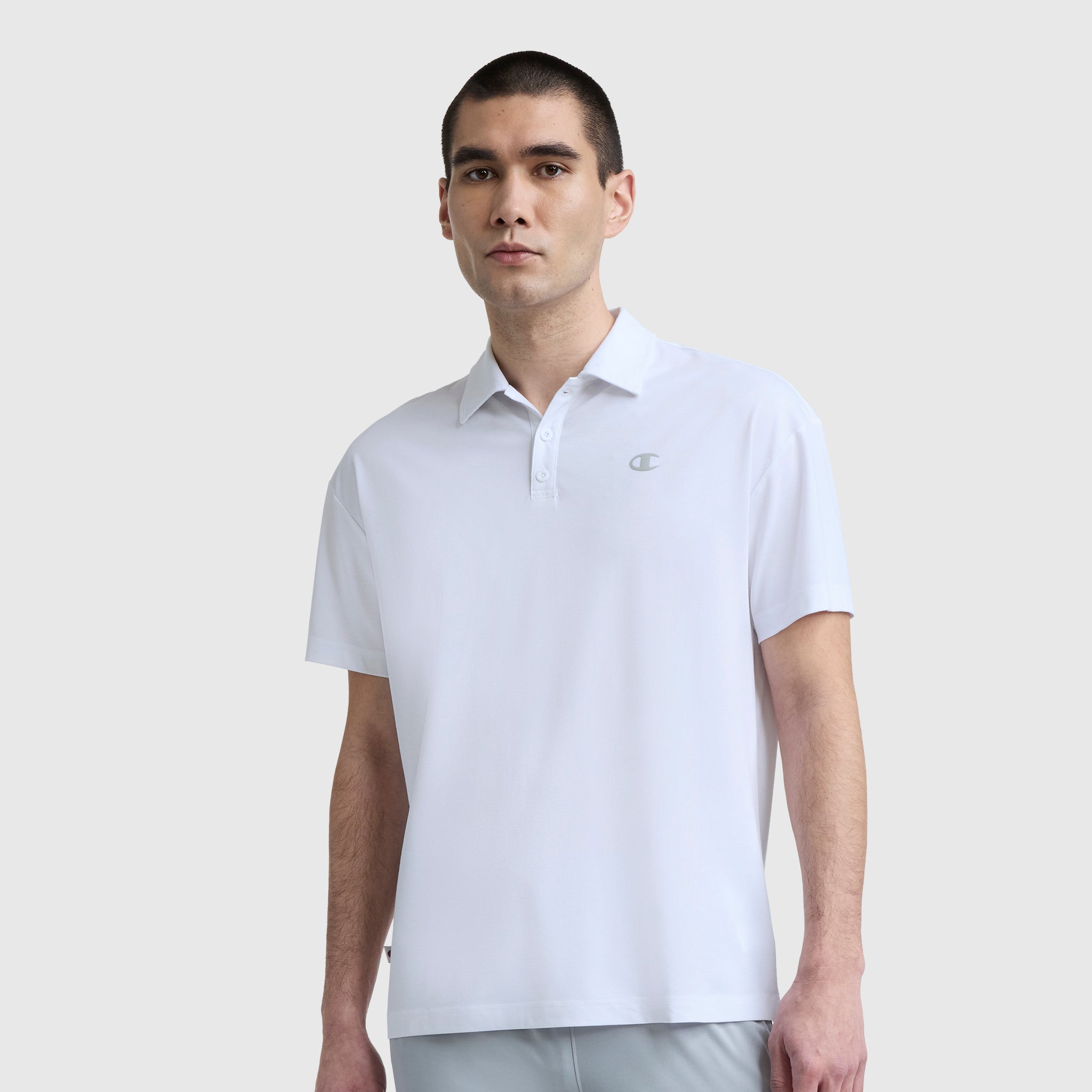 A man with short dark hair wears a plain white Champion Sport Polo, C Logo, featuring a small chest logo for an athletic look as he stands against a light gray background.