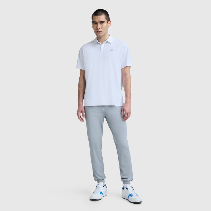 A man faces forward wearing a Champion Sport Polo, C Logo in white, light gray four-way stretch jogger pants, and white sneakers with blue accents, standing against a plain light gray background.