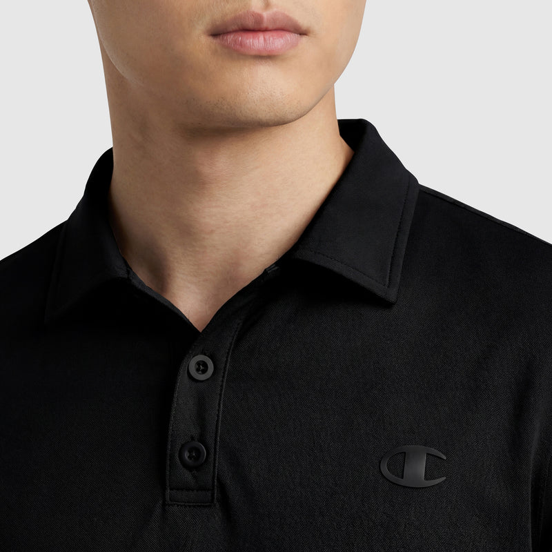 A person wears the Champion Sport Polo, C Logo—a black collared shirt with buttons and a small black Champion logo on the left chest—shown from neck to upper chest against a plain light background for an athletic look.
