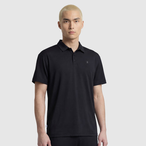 A person with short, bleached blond hair models the black Champion Sport Polo, C Logo against a plain light gray background, facing the camera with a confident athletic look.