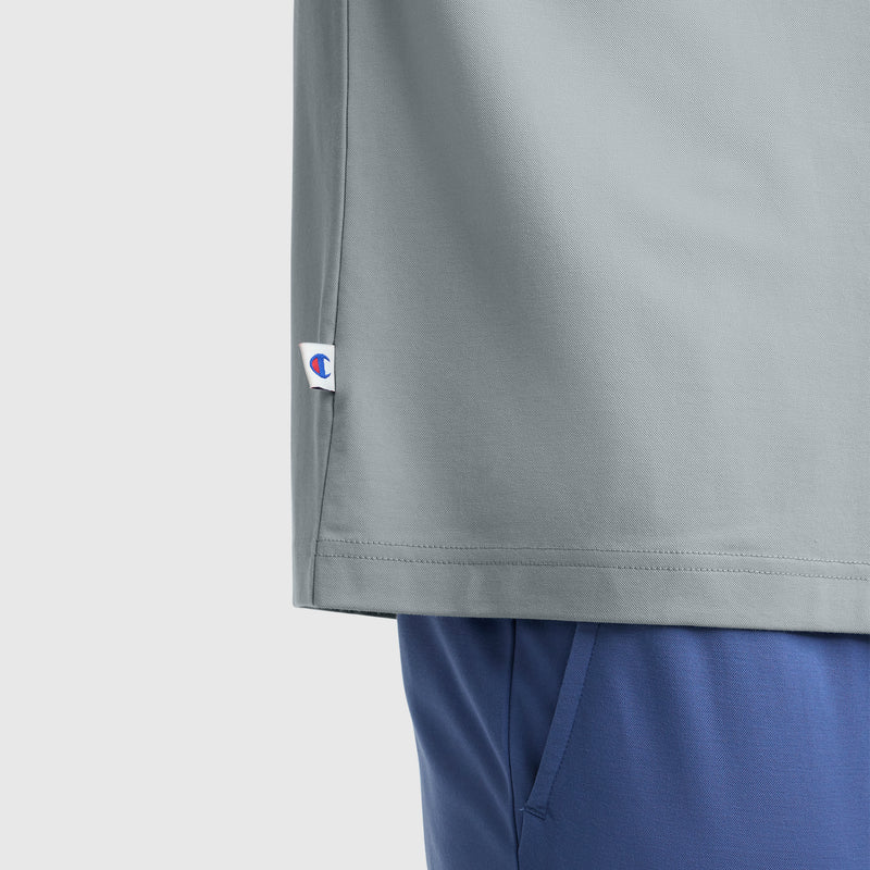 Partial view of a person wearing a light gray Champion Sport Polo with C Logo and blue pants. The Champion tag appears near the bottom hem. Background is plain and light-colored.