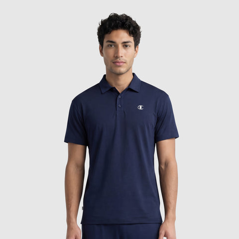A young man with short, curly dark hair models the Champion Sport Polo, C Logo in navy blue, featuring a small logo on the left chest, standing against a plain light gray background for an athletic look.