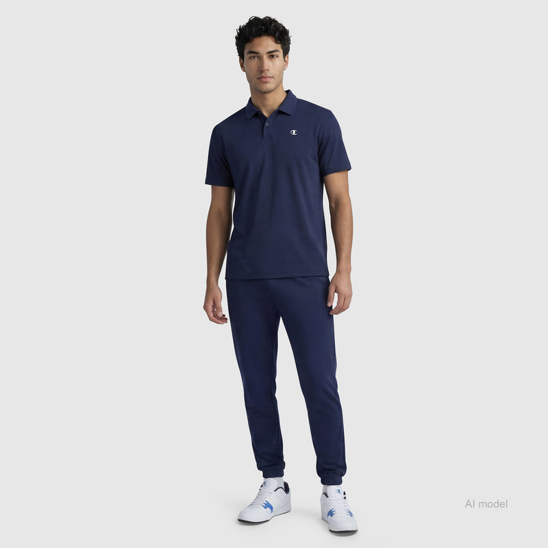 A man stands against a plain background wearing a navy blue Champion Sport Polo, C Logo, matching navy jogger pants, and white sneakers.