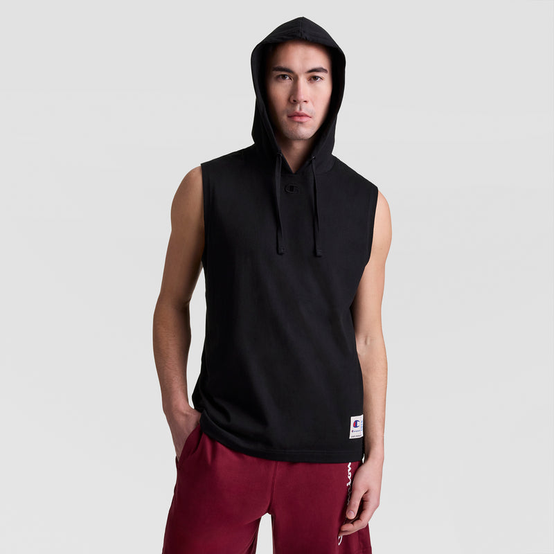 A man wearing a Champion Sleeveless Hoodie, C Logo, and maroon pants stands with one hand in his pocket against a plain light gray background.
