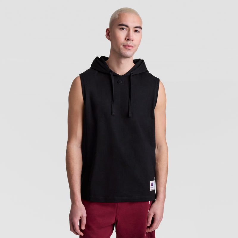A person with short blond hair wears a Champion Sleeveless Hoodie, C Logo, featuring a front pocket and drawstrings, paired with maroon shorts, standing against a plain light gray background.