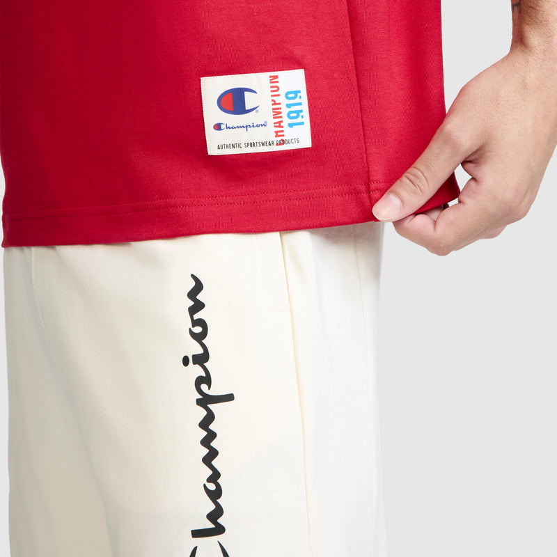 Close-up of a person wearing a red Champion Jersey Sleeveless Hoodie, featuring the C logo, made from soft ring-spun cotton with a rectangular logo tag. They are also wearing white pants with the word "Champion" written in black script along the side and are holding the hem of the hoodie.