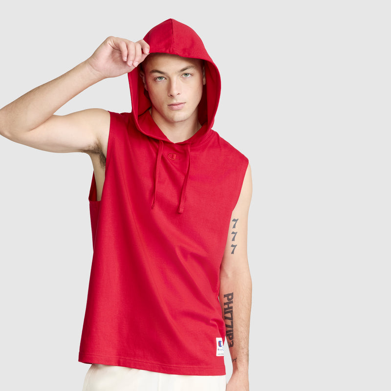 A person wearing the Champion Jersey Sleeveless Hoodie with C Logo in red, crafted from soft ring-spun cotton, and white shorts lifts the hood with one hand. Tattoos decorate their left arm as they stand against a plain light gray background.