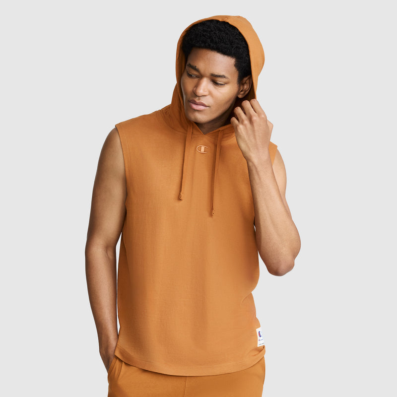 A person wearing the Champion Jersey Sleeveless Hoodie with the signature C Logo in orange, made from soft ring-spun cotton and styled with the hood up, stands against a plain light gray background, looking down while touching the side of their head.