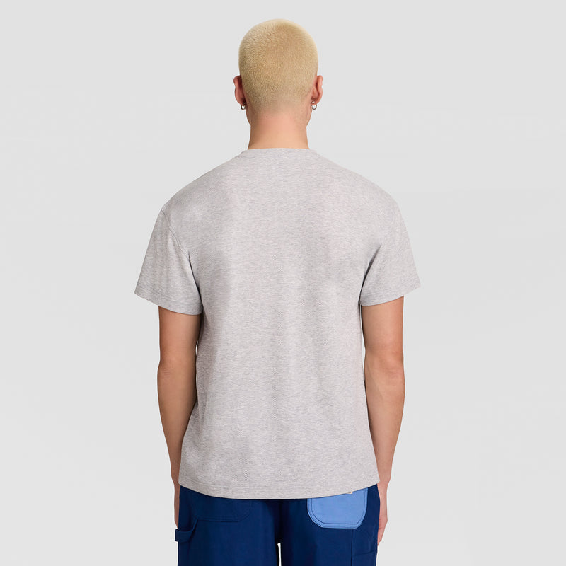 A person with short blond hair is seen from the back, wearing a light gray Champion Textured Henley with a C Logo and blue pants featuring a light blue pocket, standing against a plain light background.