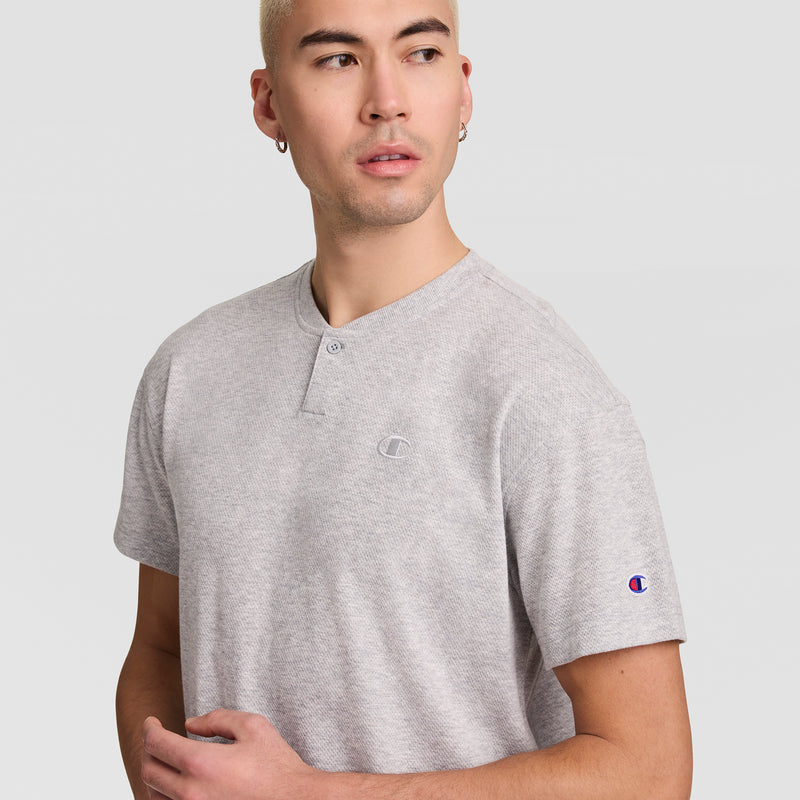 A person with short blond hair wears a light gray Champion Textured Henley, featuring the C Logo, and looks to the side with a neutral expression against a plain light background.
