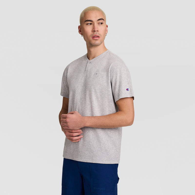A person with short blond hair is wearing the Champion Textured Henley, C Logo top in light gray and blue pants, standing against a plain light background while looking slightly to the side.