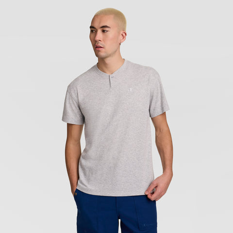 A person with short blond hair is wearing the Champion Textured Henley, C Logo in light gray, styled with blue pants. They stand against a plain light gray background, looking slightly to the side.