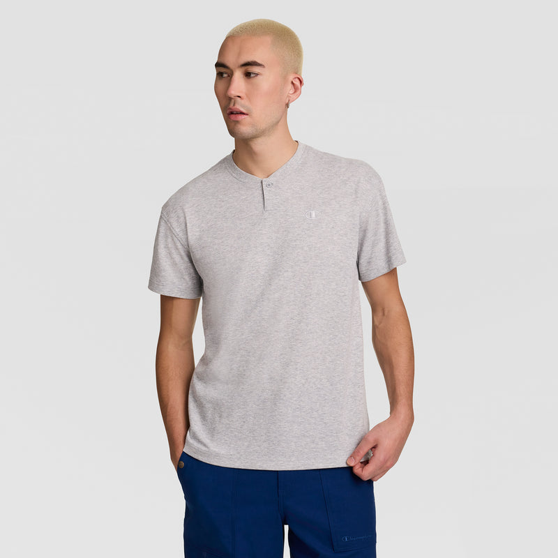 A person with short blond hair is wearing the Champion Textured Henley, C Logo in light gray, styled with blue pants. They stand against a plain light gray background, looking slightly to the side.