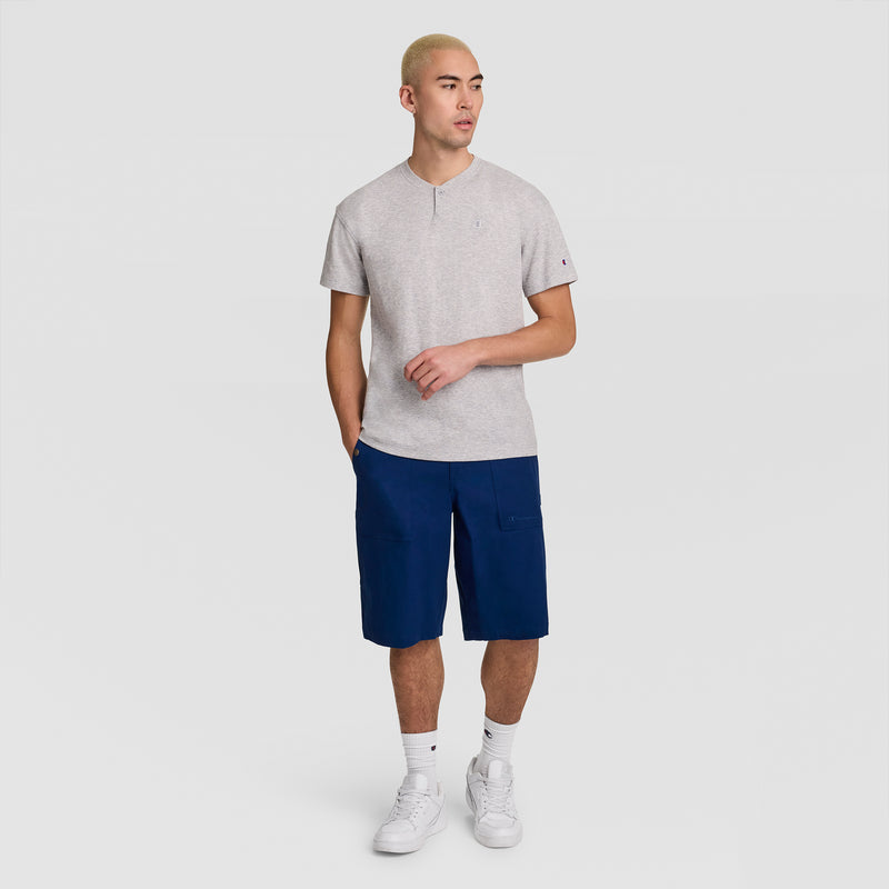 A person with short blond hair wears a Champion Textured Henley, C Logo styled as a men’s standard-fit top, along with navy blue shorts, white socks, and white sneakers, standing against a plain light gray background.