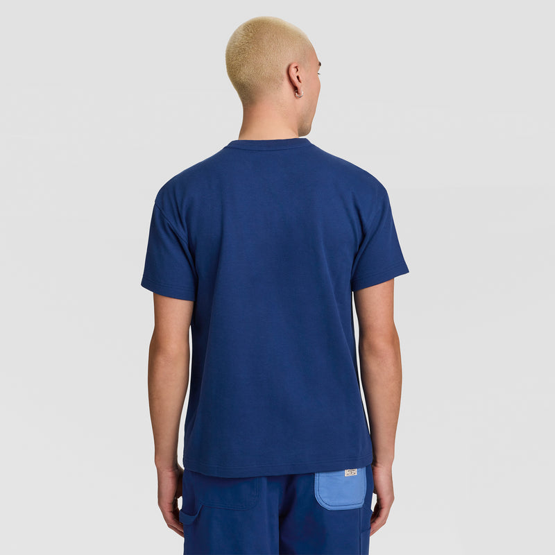 A person with short blond hair is standing with their back to the camera against a plain background, wearing a navy blue Champion Textured Henley, C Logo men’s standard-fit top and blue pants featuring a light blue back pocket.
