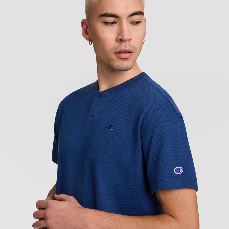 A person with short blond hair wearing a blue Champion Textured Henley, C Logo top looks to the side. The background is plain light gray.