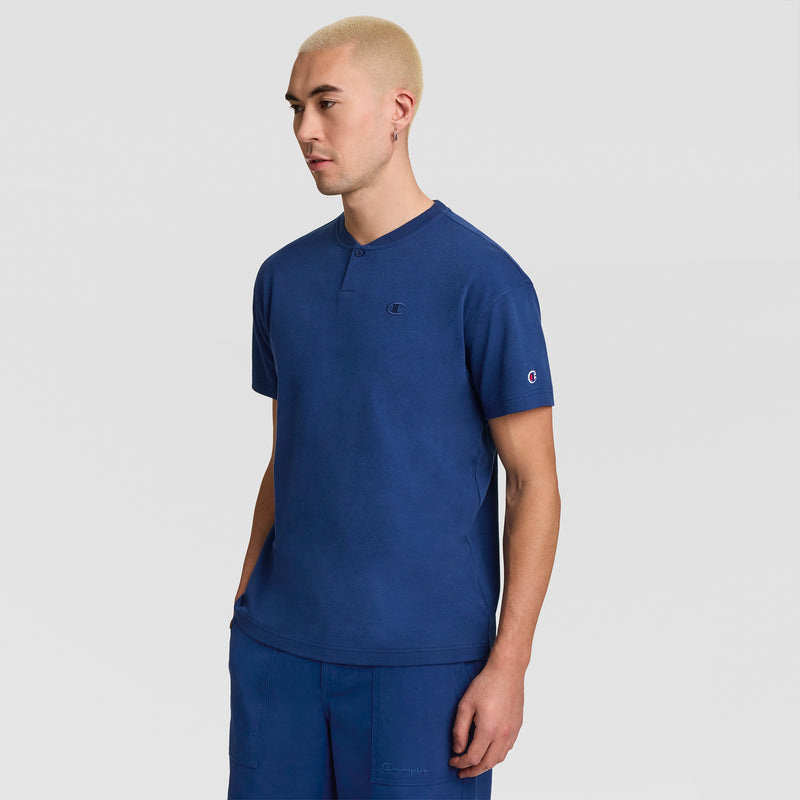 A man with short blond hair is wearing a blue Champion Textured Henley, C Logo shirt paired with matching blue shorts, standing against a plain light gray background.