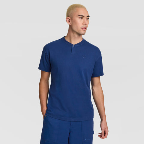 A person with short blonde hair wears a blue Champion Textured Henley, C Logo shirt and matching blue pants, standing against a plain light gray background.