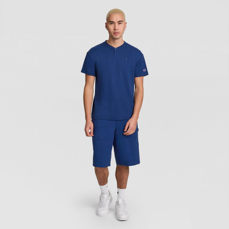 A person with short blond hair wears the Champion Textured Henley, C Logo men’s standard-fit top in blue and matching blue shorts, paired with white socks and Champion white sneakers, standing against a plain white background.