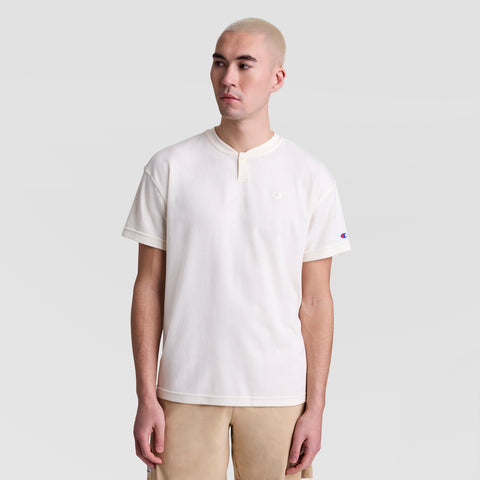 A person with short, light hair wears the Champion Textured Henley with a tonal embroidered C logo and buttoned collar, standing against a plain light gray background.
