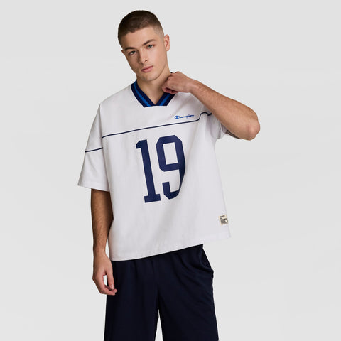 A young man in vintage style stands against a light gray backdrop, wearing a loose white Champion Gridiron Football Jersey with script logo and number 19 in navy, paired with dark shorts, looking at the camera while touching his neck.