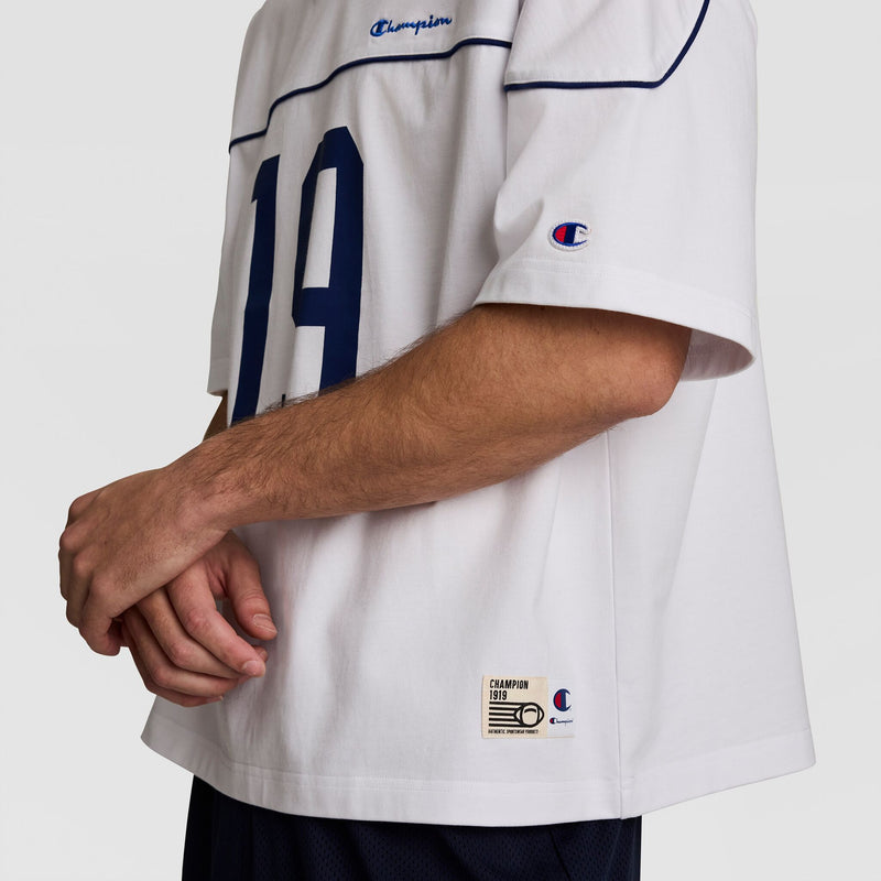 A person wears the Champion Gridiron Football Jersey, Script Logo—a white jersey with blue "19," Champion logos on the chest and sleeve, and a stitched hem label—showcasing vintage gridiron style as they cross their arms at the waist.