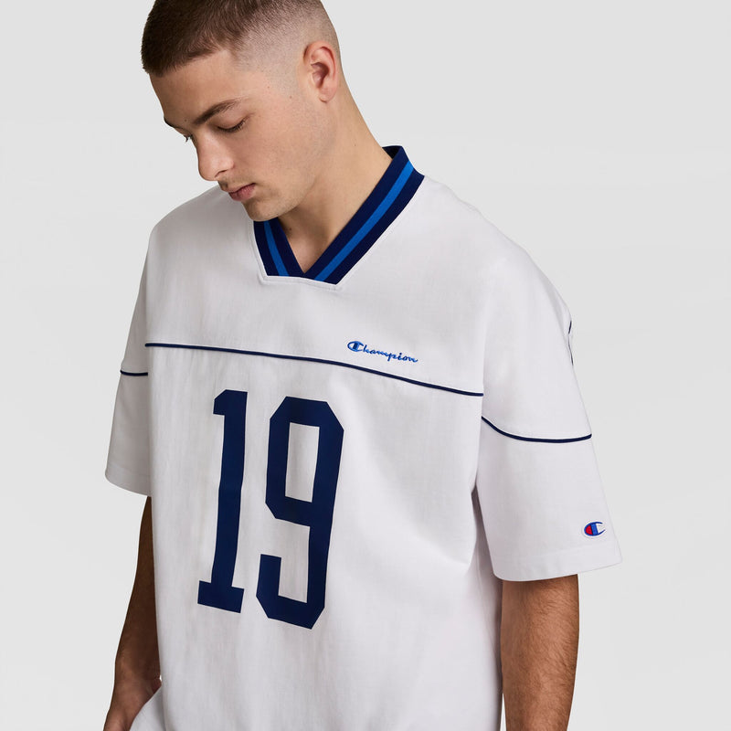 A young man with short hair looks down while wearing the Champion Gridiron Football Jersey, Script Logo. The vintage style features blue trim and the number 19, with Champion logos on the chest and sleeve against a plain light gray background.