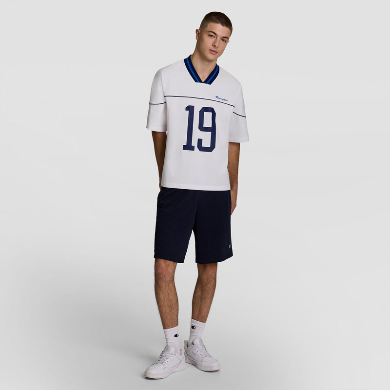 A young man stands against a plain backdrop, wearing the Champion Gridiron Football Jersey with script logo, featuring a blue collar and number 19, paired with navy shorts, white socks, and vintage-style white sneakers.