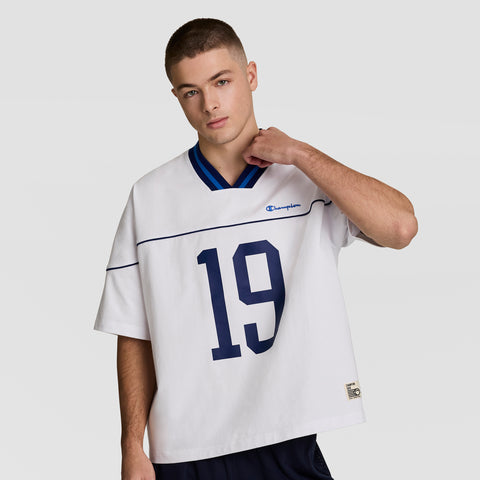 A young man with short hair wears a loose white Champion Gridiron Football Jersey, Script Logo, featuring the number 19 in navy blue, showcasing vintage athletic style against a plain light gray background.