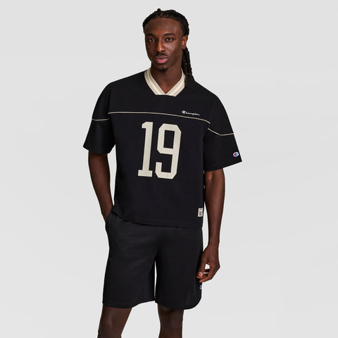 A man with long braids wears a black short-sleeve jersey with the number 19 on the front and matching black shorts, standing against a plain light gray background.