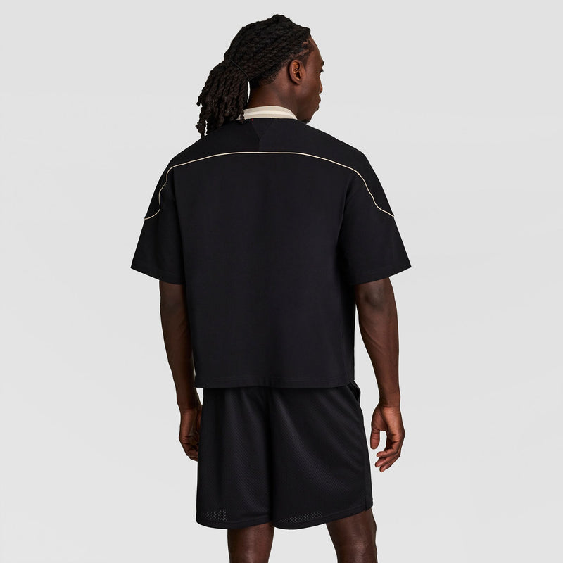 A man with braided hair wearing a black short-sleeve shirt with white piping detail and black athletic shorts stands facing away from the camera against a plain light background.