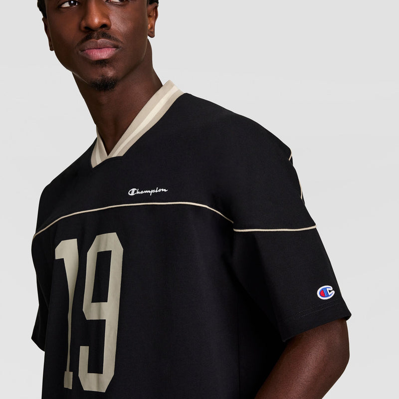 A man wearing a black Champion jersey with beige trim and the number 19 on the front. The Champion logo appears on the chest and sleeve. The background is plain light gray.
