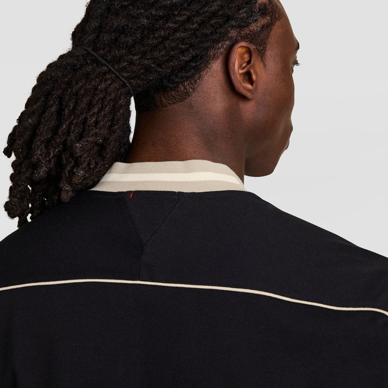 A person with long, dark dreadlocks tied back is shown from behind, wearing a black shirt with a light-colored collar and a thin, light trim across the back.