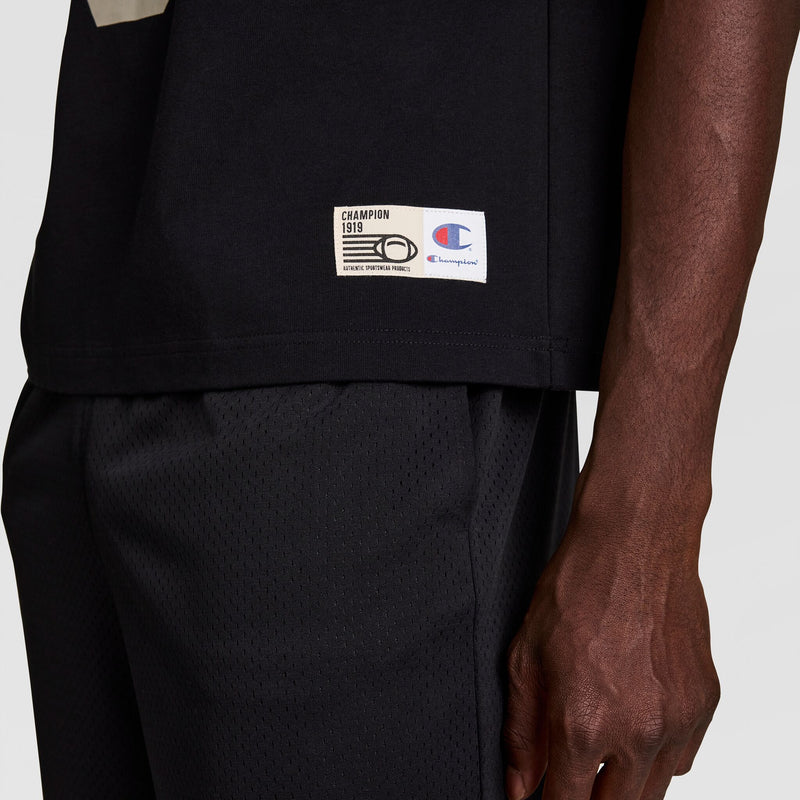 A close-up of a person wearing black mesh athletic shorts and a black Champion t-shirt. The Champion logo and label are visible near the shirt hem, and the persons hand is relaxed at their side.