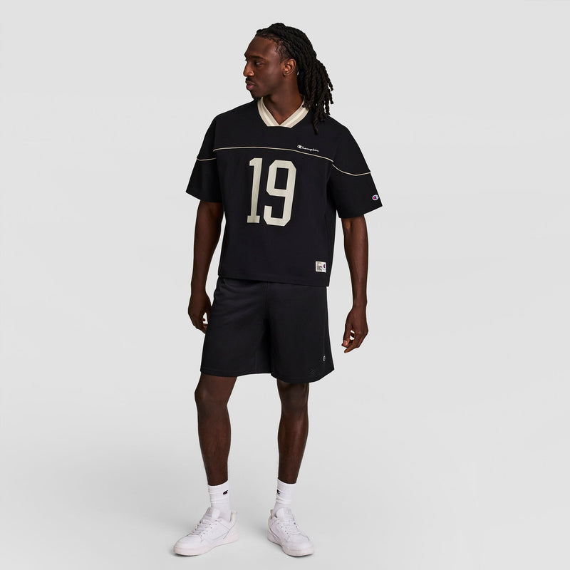 A man stands wearing a black sports jersey with the number 19, black shorts, and white sneakers, looking to his left against a plain light gray background.