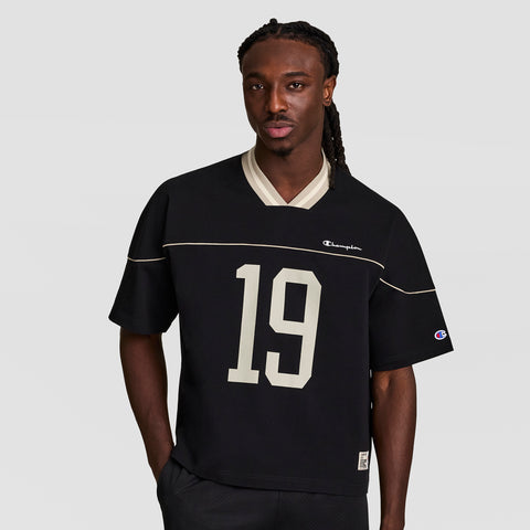 A man with long braided hair stands against a light gray background wearing the Champion Gridiron Football Jersey, Script Logo, in black with large white number 19—showcasing vintage athletic style and casual confidence.