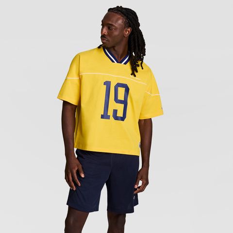 A person with long braids wears a loose yellow Gridiron Football Jersey by Champion featuring the script logo and number 19 in navy blue, along with navy blue shorts, standing against a plain light gray background.