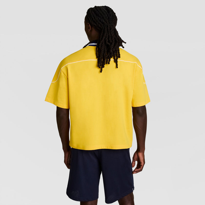 With their back to the camera, a person with long dark dreadlocks showcases vintage athletic style in a loose-fitting yellow Champion Gridiron Football Jersey, Script Logo, paired with navy shorts against a plain light gray background.