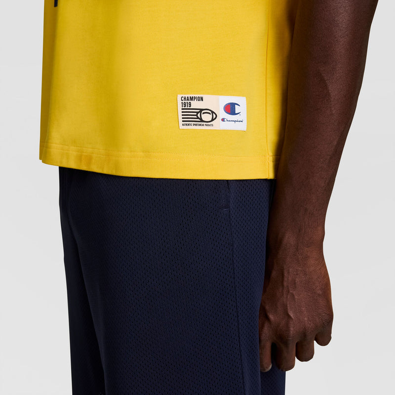 A person wearing a yellow Champion Gridiron Football Jersey, Script Logo, in vintage athletic style and dark blue shorts stands with their arm relaxed. The Champion script logo and tag appear at the bottom of the shirt.