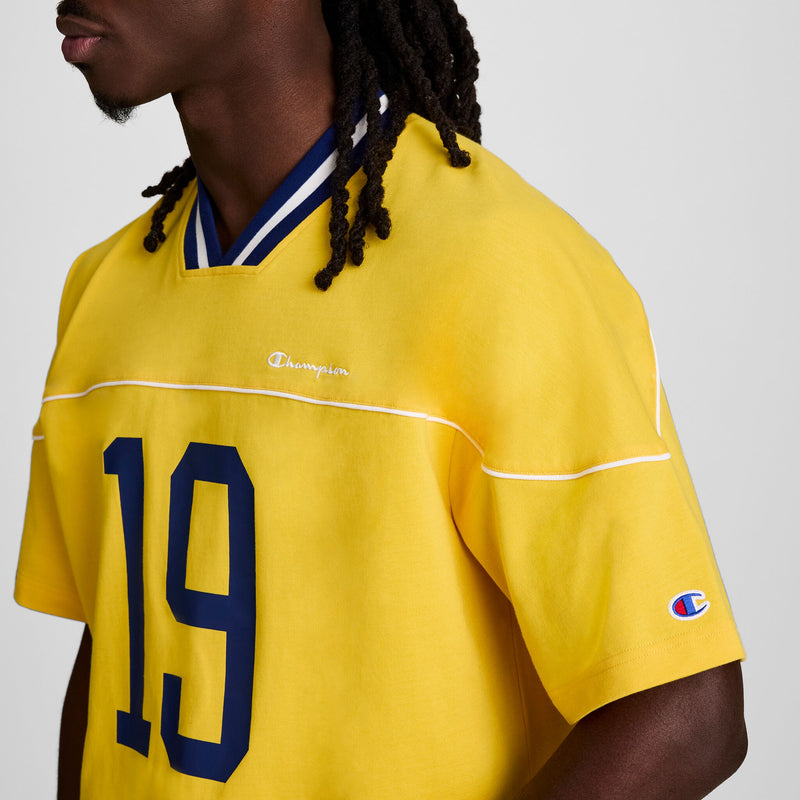 A person with long braids is wearing a yellow Champion Gridiron Football Jersey, Script Logo, featuring blue trim and the number 19. The photo shows their upper body facing left.