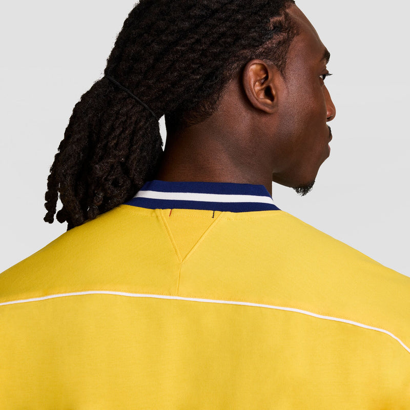 Seen from behind, a person with dark braided hair wears the Champion Gridiron Football Jersey, Script Logo—featuring yellow fabric, white piping, and a blue-and-white striped collar—showcasing vintage athletic style against a plain gray background.