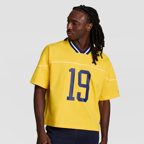 A man with long braided hair wears a bright yellow Champion Gridiron Football Jersey, Script Logo, with a blue collar and bold blue 19, in vintage athletic style. He looks left against a plain light background.