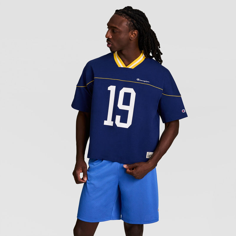 A man with long braided hair wears the Champion Gridiron Football Jersey, Script Logo—blue with number 19 and yellow trim—paired with blue athletic shorts, standing slightly to the side against a plain gray background for a vintage athletic look.