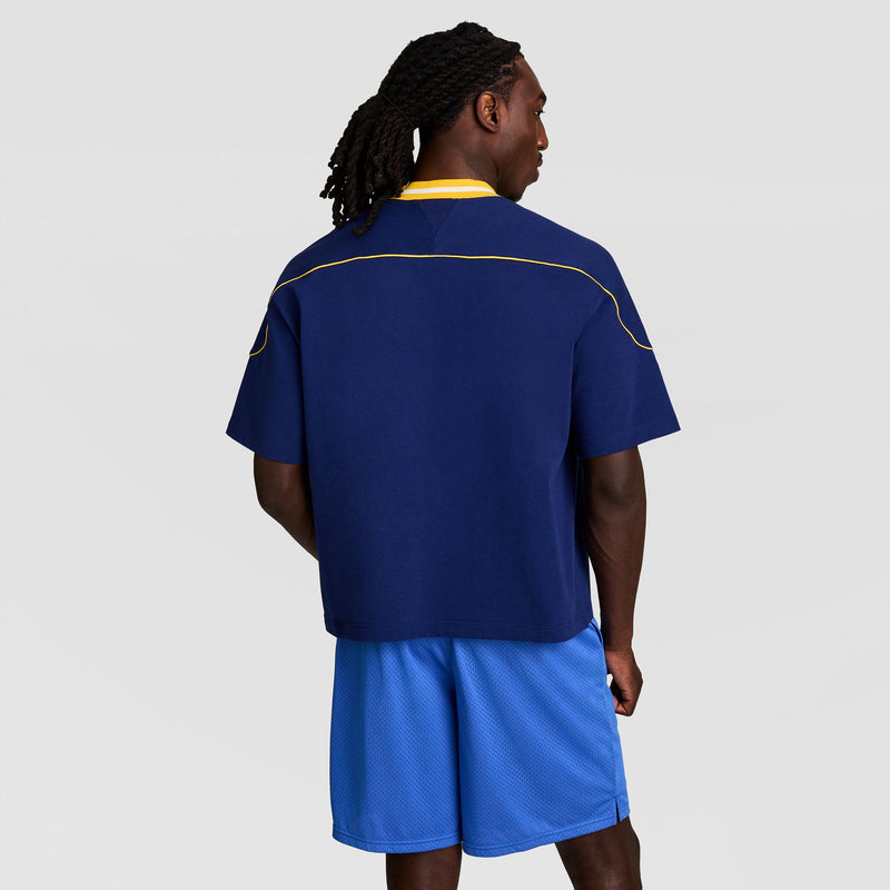 A man with long, braided hair faces away, wearing the Champion Gridiron Football Jersey Script Logo in navy blue with yellow piping and light blue shorts, channeling a vintage athletic style against a plain light gray background.